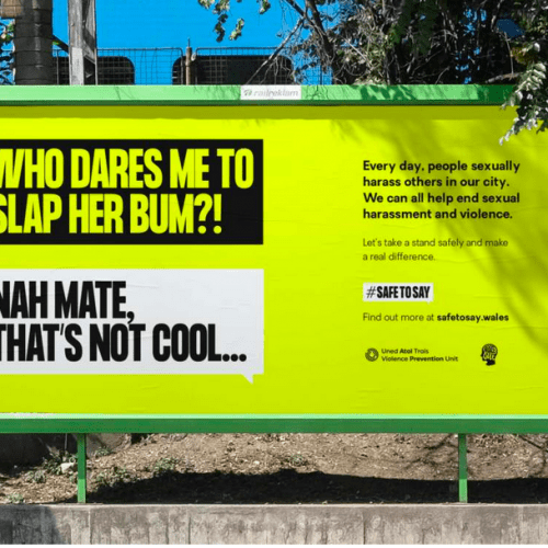 A billboard in Cardiff. The poster depicts a conversation between two people via speech bubbles. The first person says, 'Who dares me to slap her bum?!' to which the second person respons 'Nah mate, that's not cool'. Adjacent text reads 'Every day, people sexually harass others in our city. We can all help end sexual harrassment and violence. Let's take a stand safely and make a real difference.' Underneath is the hashtag #SafeToSay and the text 'Find out more at safetosay.wales.'